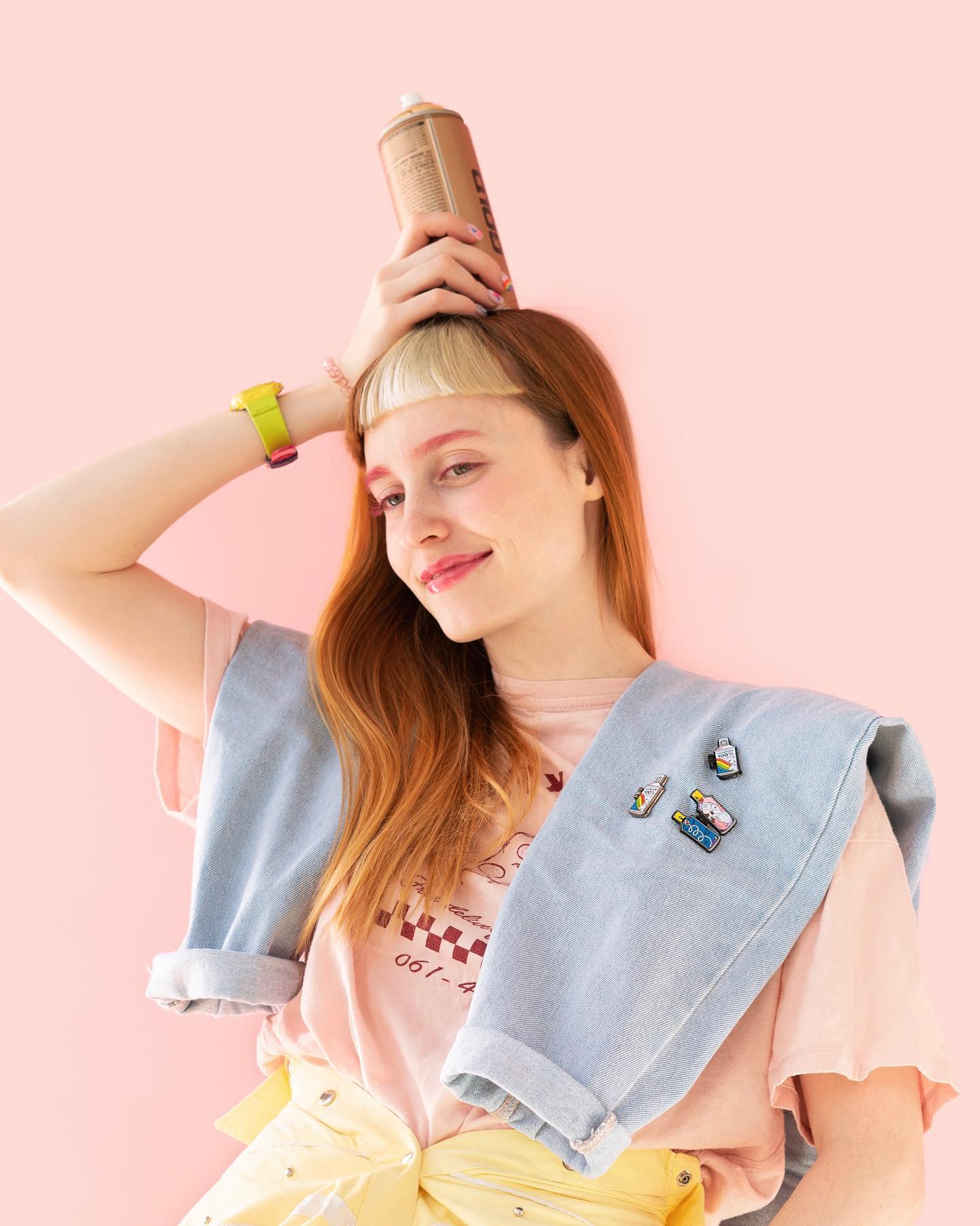 Person holding a spray can on their head while wearing pastel clothing and decorative enamel pins against a pink background.