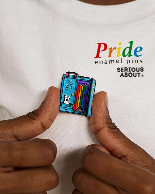 Hands holding a rainbow-themed enamel pin shaped like a locker on a white T-shirt that reads “Pride enamel pins.”