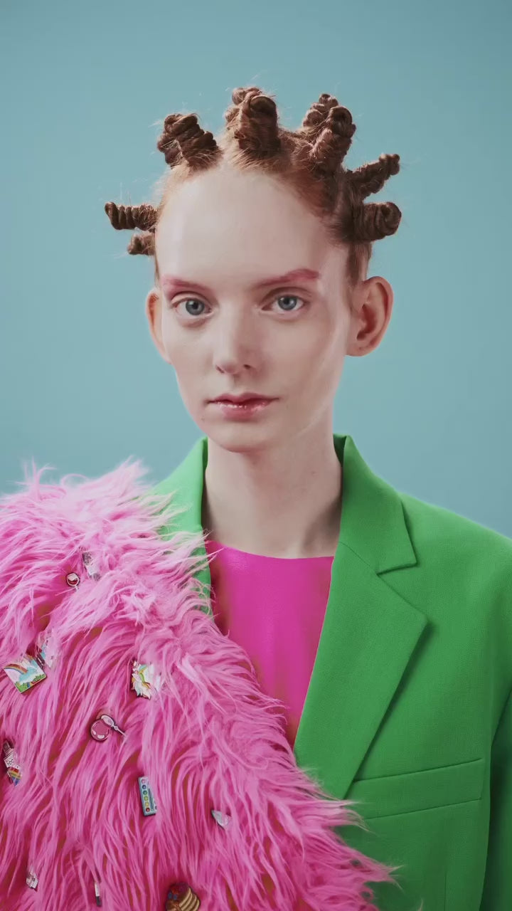 Close-up portrait of a person with small twisted buns hairstyle, dressed in a bright green blazer and pink textured layer with decorative pins.