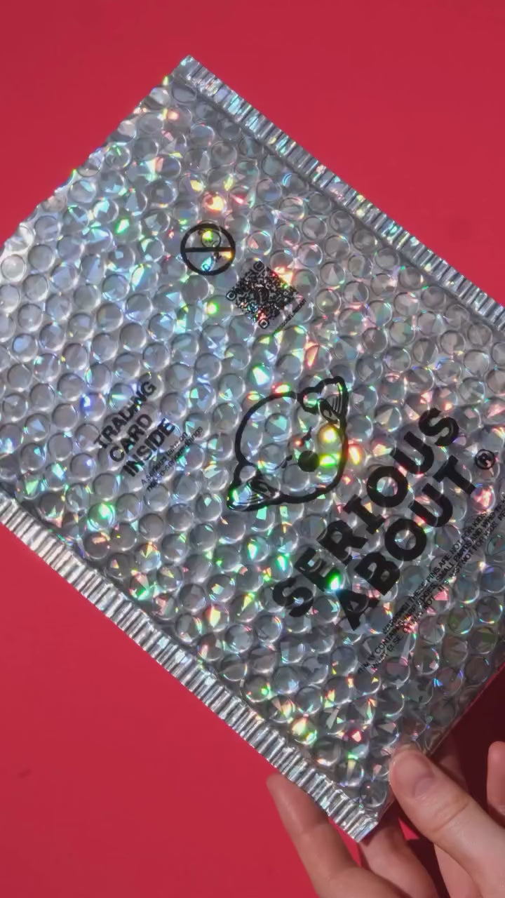Close-up of a silver holographic bubble mailer with “Serious About” logo and “Trading card inside” text on a red backdrop.
