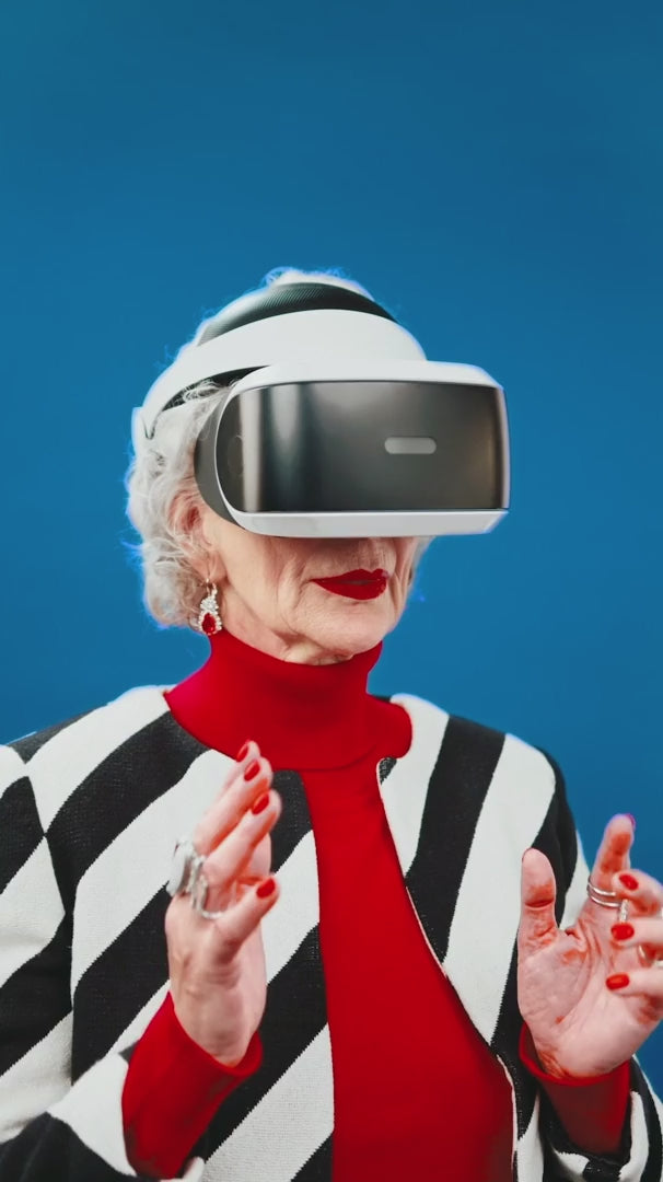 Portrait of an older woman in a red turtleneck and striped jacket using a virtual reality headset against a blue backdrop.