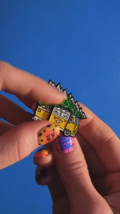 Close-up of hands displaying a yellow camper van enamel pin decorated with green trees on a bright blue backdrop.