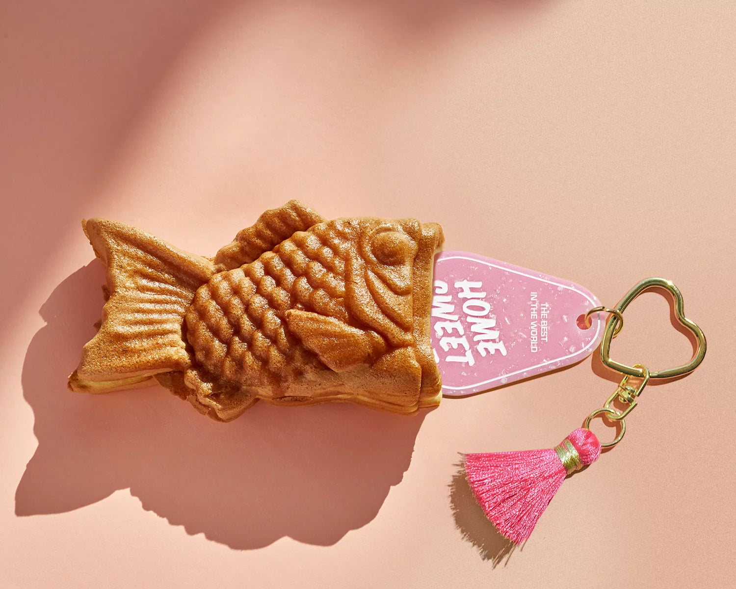 Taiyaki-style fish waffle keychain with a pink “Home Sweet Home” tag, gold heart clasp, and pink tassel on a peach backdrop.